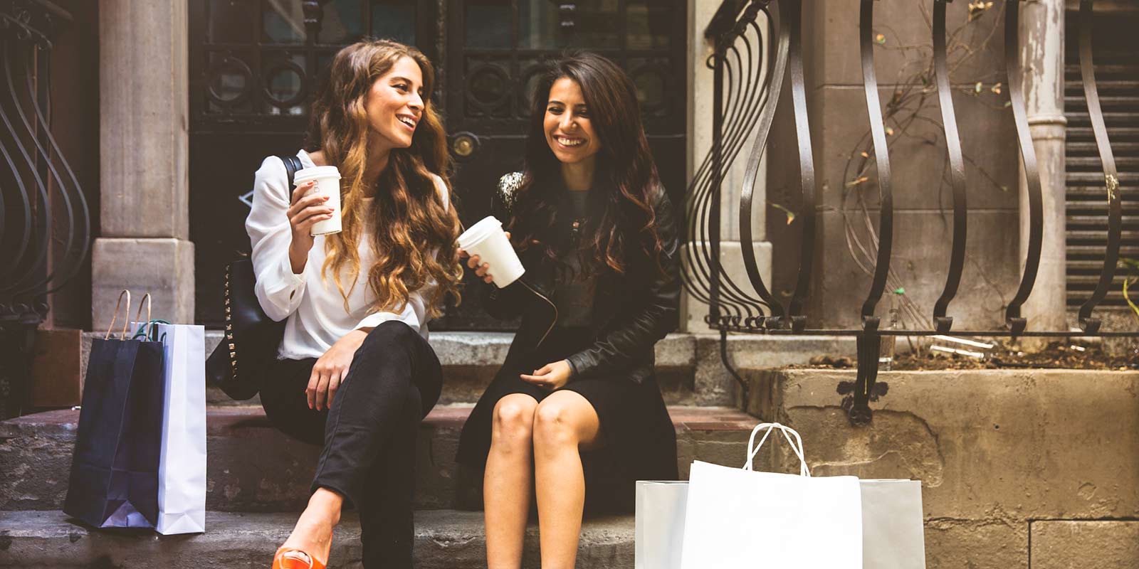 Two women sit on steps and enjoy coffee together after a day of shopping in the city.